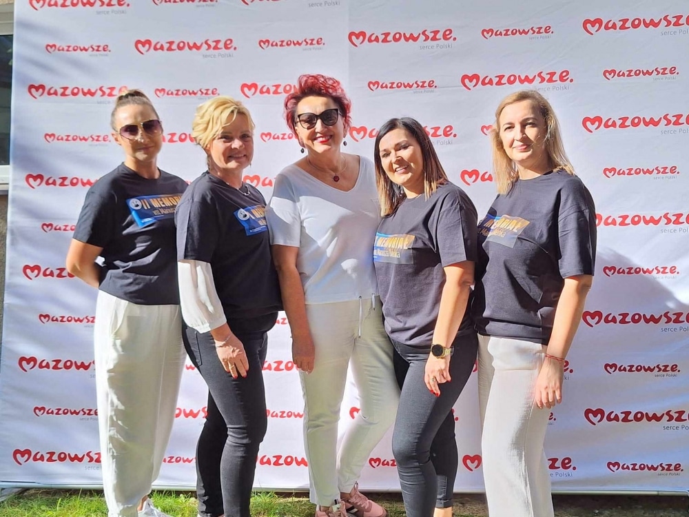 Grupa pięciu kobiet w różnych ubraniach, stojących na tle z logo Mazowsza, pozuje do zdjęcia na wydarzeniu lokalnym.