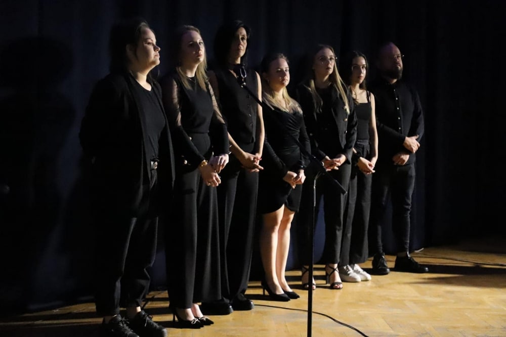 Na zdjęciu widać grupę sześciu osób w ciemnych strojach, stojących w szeregu na scenie, na tle ciemnego materiału.