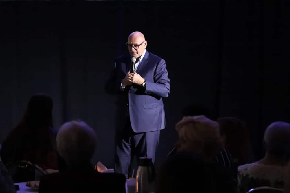 Na zdjęciu widoczny jest mężczyzna w garniturze, stojący na scenie z mikrofonem w ręku, podczas wydarzenia, z uczestnikami siedzącymi w tle.