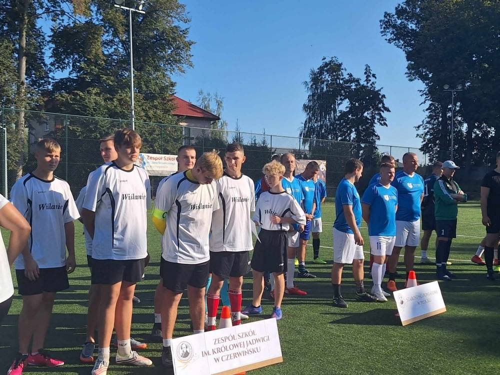Uczestnicy turnieju piłkarskiego stoją w kolejkach przed meczem na boisku, prezentując różne stroje sportowe.