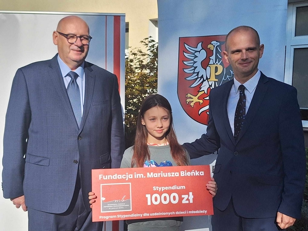 Na zdjęciu trzech ludzi stoi obok siebie, a dziewczynka trzyma wielki czek z napisem „Fundacja im. Mariusza Bieńka, Stypendium 1000 zł”.