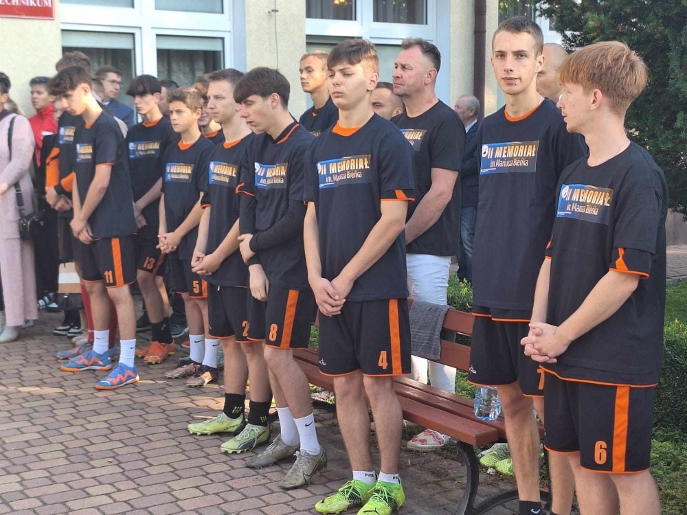 Na zdjęciu widać grupę młodych sportowców w koszulkach z napisem „III MEMORIAŁ im. Mariusza Bieka”, którzy stoją w serii na zewnątrz budynku.