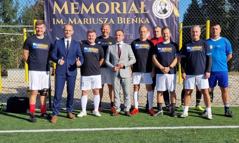 Na zdjęciu przedstawiono grupę mężczyzn w strojach sportowych, którzy pozują przed bannerem z napisem "Memoriał im. Mariusza Bieńka" na boisku piłkarskim.