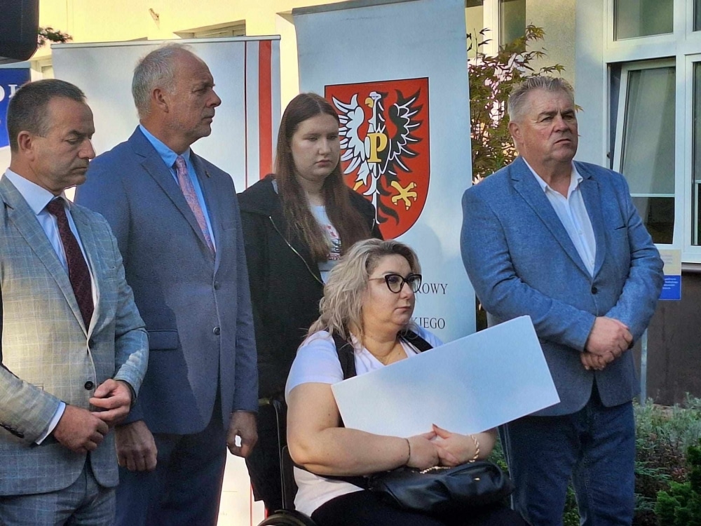 Uczestnicy wydarzenia stoją w grupie przed budynkiem, gdzie widoczne są banner z herbem oraz różne elementy reklamy.