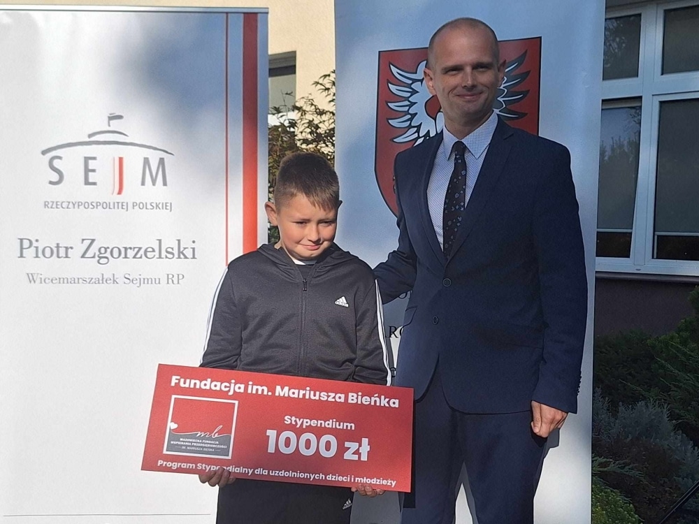 Chłopiec w dresie stoi obok mężczyzny w garniturze, trzymając dużą tabliczkę z napisem "Stypendium 1000 zł" w tle z logo Sejmu i nazwiskiem Piotra Zgorzelskiego.