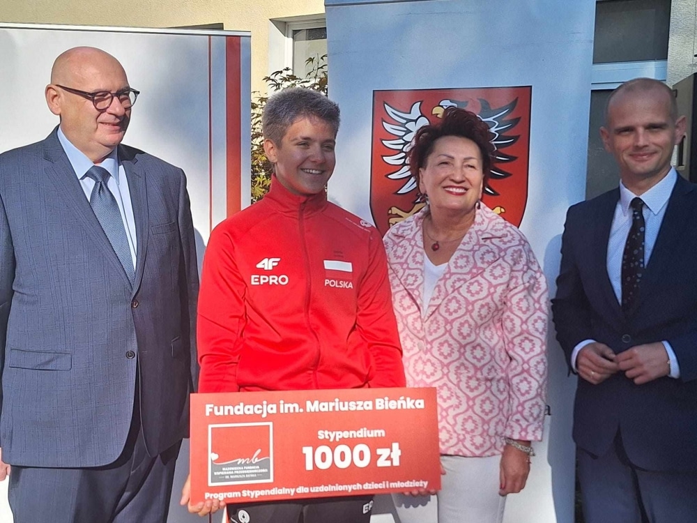 Na zdjęciu czterech ludzi stoi obok siebie, trzymając czek w wysokości 1000 złotych, symbolizujący stypendium Fundacji im. Mariusza Bieńka.