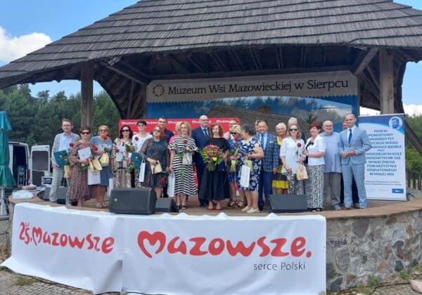 Na zdjęciu grupa osób stoi na scenie przed Muzeum Wsi Mazowieckiej w Sierpcu, trzymając kwiaty i dyplomy, celebrując wydarzenie związane z 25-leciem Mazowsza.