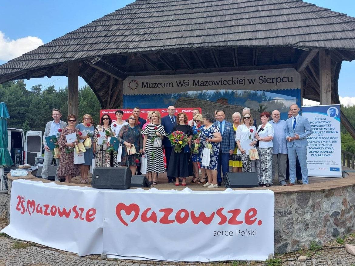 Na zdjęciu grupa osób stoi na scenie przed Muzeum Wsi Mazowieckiej w Sierpcu, trzymając kwiaty i dyplomy, celebrując wydarzenie związane z 25-leciem Mazowsza.