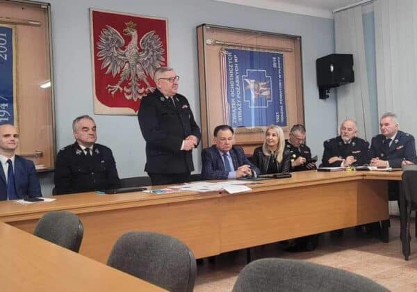 Zdjęcie przedstawia grupę osób w mundurach zaproszonych na spotkanie, siedzących przy stole w sali konferencyjnej, z emblemata­mi i flagami w tle.