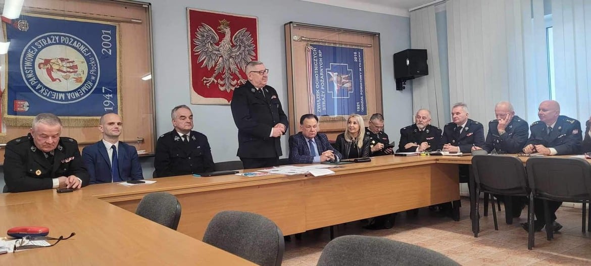 Zdjęcie przedstawia grupę osób w mundurach zaproszonych na spotkanie, siedzących przy stole w sali konferencyjnej, z emblemata­mi i flagami w tle.