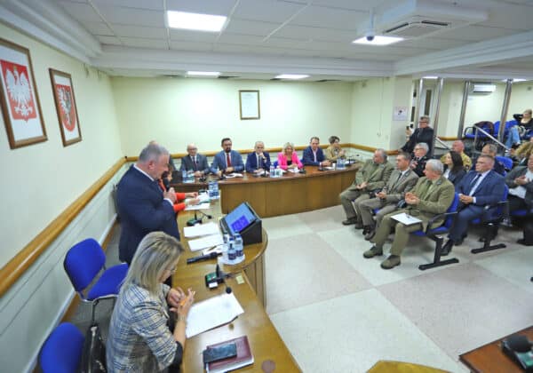 Na zdjęciu przedstawiono zebranie, podczas którego przewodniczący przemawia do zgromadzonych uczestników siedzących przy stole.