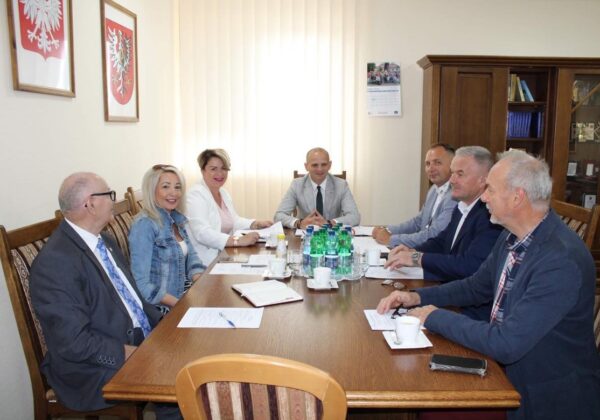 Na zdjęciu przedstawiono grupę siedmiu osób w formalnym otoczeniu, uczestniczących w spotkaniu przy stole, z dokumentami i napojami na stole.