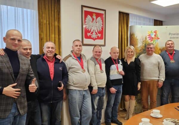 Na zdjęciu przedstawiono grupę mężczyzn z medalami oraz kobietę, wszyscy ustawieni w sali z widokiem na flagę i baner Powiatu Płockiego.