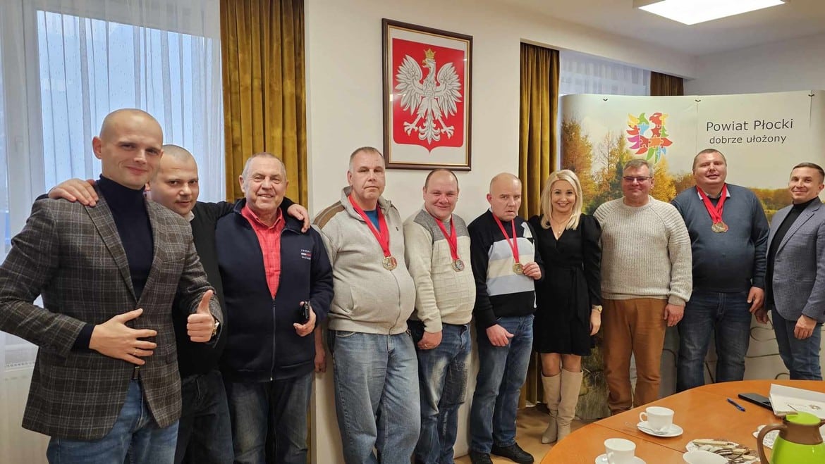 Na zdjęciu przedstawiono grupę mężczyzn z medalami oraz kobietę, wszyscy ustawieni w sali z widokiem na flagę i baner Powiatu Płockiego.