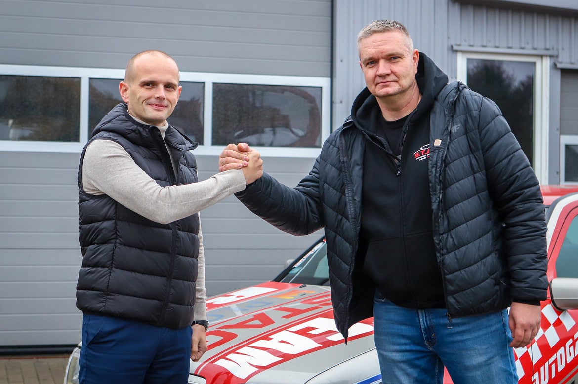 Na zdjęciu dwóch mężczyzn uścisnęło sobie ręce na tle samochodu i szarej budynku.