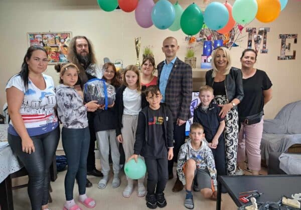 Grupa dzieci i dorosłych stoi w radosnej atmosferze przed dekoracjami urodzinowymi z balonami, trzymając podarunek.