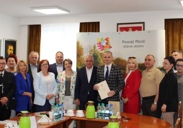 Na zdjęciu grupa osób stoi w sali, trzymając dokumenty, a w tle widoczny jest baner z logo Powiatu Płockiego.