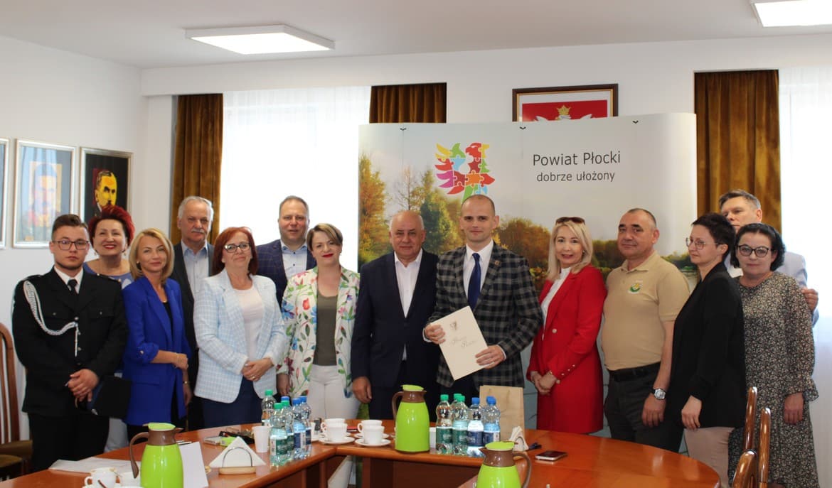 Na zdjęciu grupa osób stoi w sali, trzymając dokumenty, a w tle widoczny jest baner z logo Powiatu Płockiego.