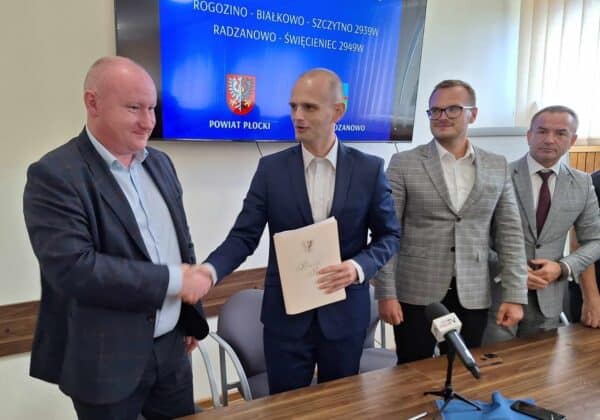 Na zdjęciu przedstawiono uroczystość podpisania umowy w obecności kilku mężczyzn, z których jeden w garniturze podaje rękę drugiemu trzymającemu dokument.