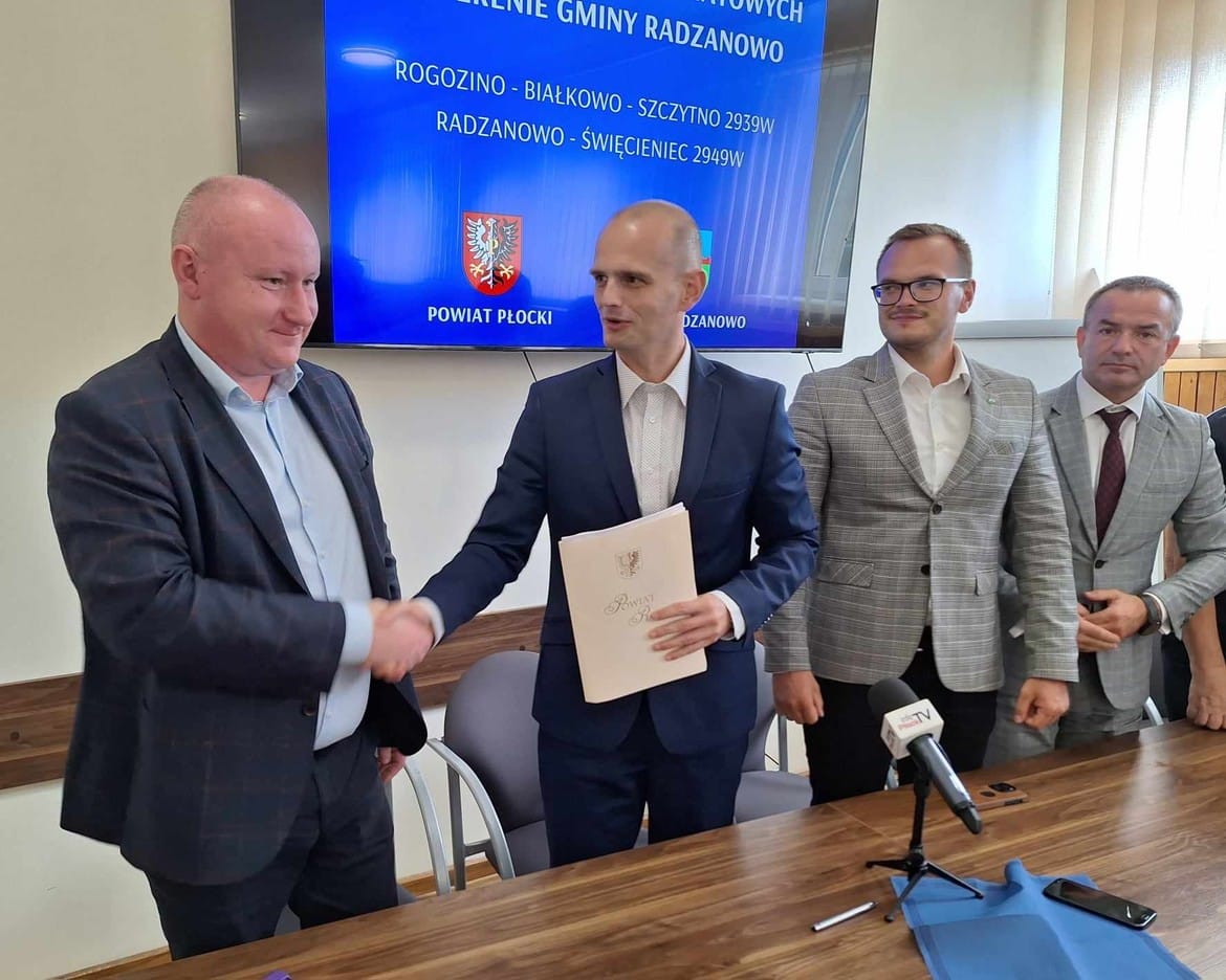 Na zdjęciu przedstawiono uroczystość podpisania umowy w obecności kilku mężczyzn, z których jeden w garniturze podaje rękę drugiemu trzymającemu dokument.