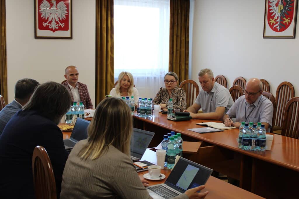 Na zdjęciu przedstawiona jest grupa ludzi w trakcie spotkania przy stole w sali konferencyjnej, z widocznymi laptopami i wodą w butelkach.