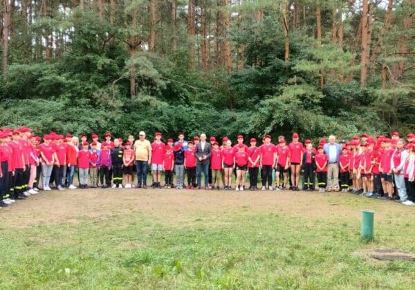 Na zdjęciu widać dużą grupę osób w czerwonych t-shirtach i czapkach, stojących w okręgu na tle lasu.