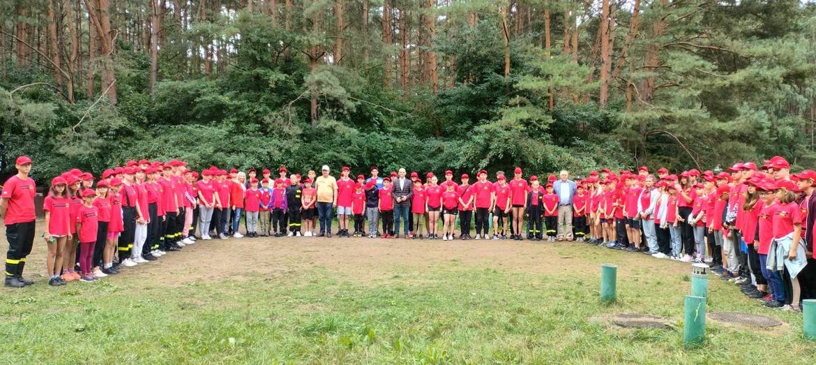 Na zdjęciu widać dużą grupę osób w czerwonych t-shirtach i czapkach, stojących w okręgu na tle lasu.