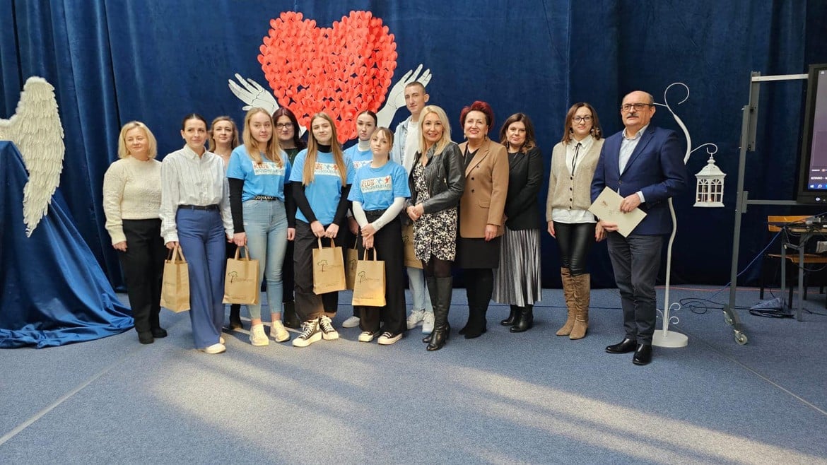 Zdjęcie przedstawia grupę osób pozujących na tle serca wykonanego z papierowych serc, w ramach wydarzenia społecznego.