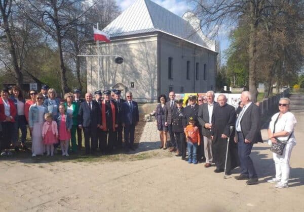 Grupa ludzi w różnym wieku pozująca przed budynkiem z flagą Polski w tle, w czasie słonecznego dnia.