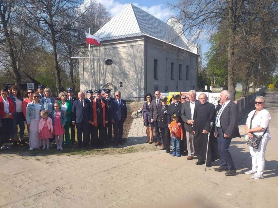 Grupa ludzi w różnym wieku pozująca przed budynkiem z flagą Polski w tle, w czasie słonecznego dnia.