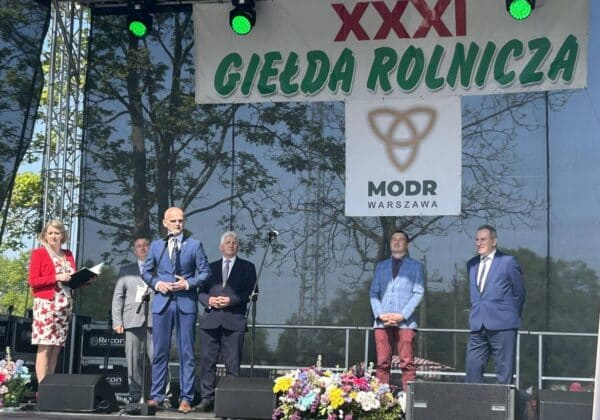 Na scenie podczas XXXI Giełdy Rolniczej stoi grupa ludzi, w tym mężczyzna przemawiający, otoczona kwiatami i banerem z logo MODR Warszawa.