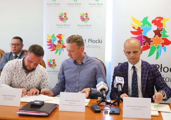 Na zdjęciu widać trzech mężczyzn siedzących przy stole, podpisujących dokumenty podczas konferencji dotyczącej powiatu płockiego.