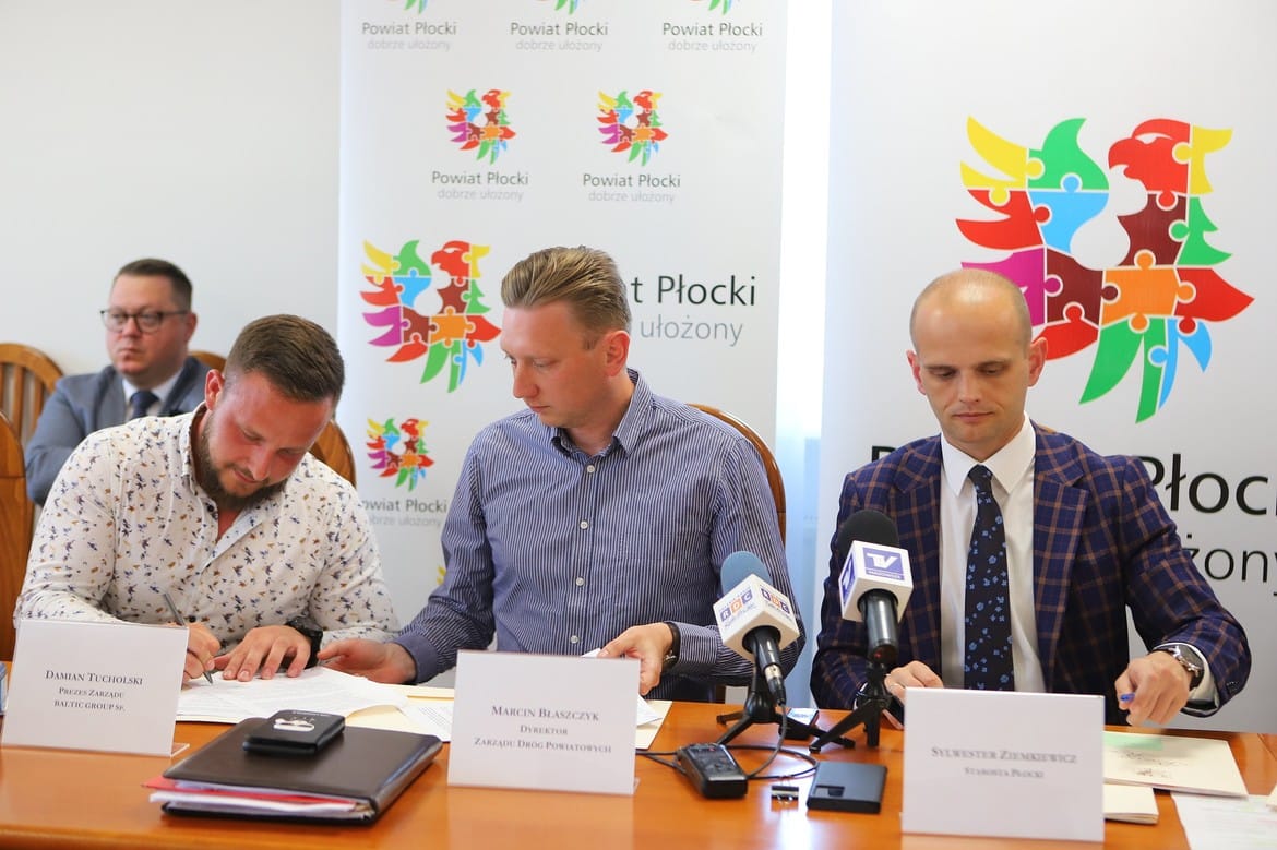 Na zdjęciu widać trzech mężczyzn siedzących przy stole, podpisujących dokumenty podczas konferencji dotyczącej powiatu płockiego.