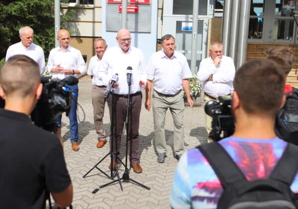 Na zdjęciu widać grupę mężczyzn w białych koszulach, którzy prowadzą konferencję prasową na zewnątrz budynku, otoczeni reporterami z kamerami.