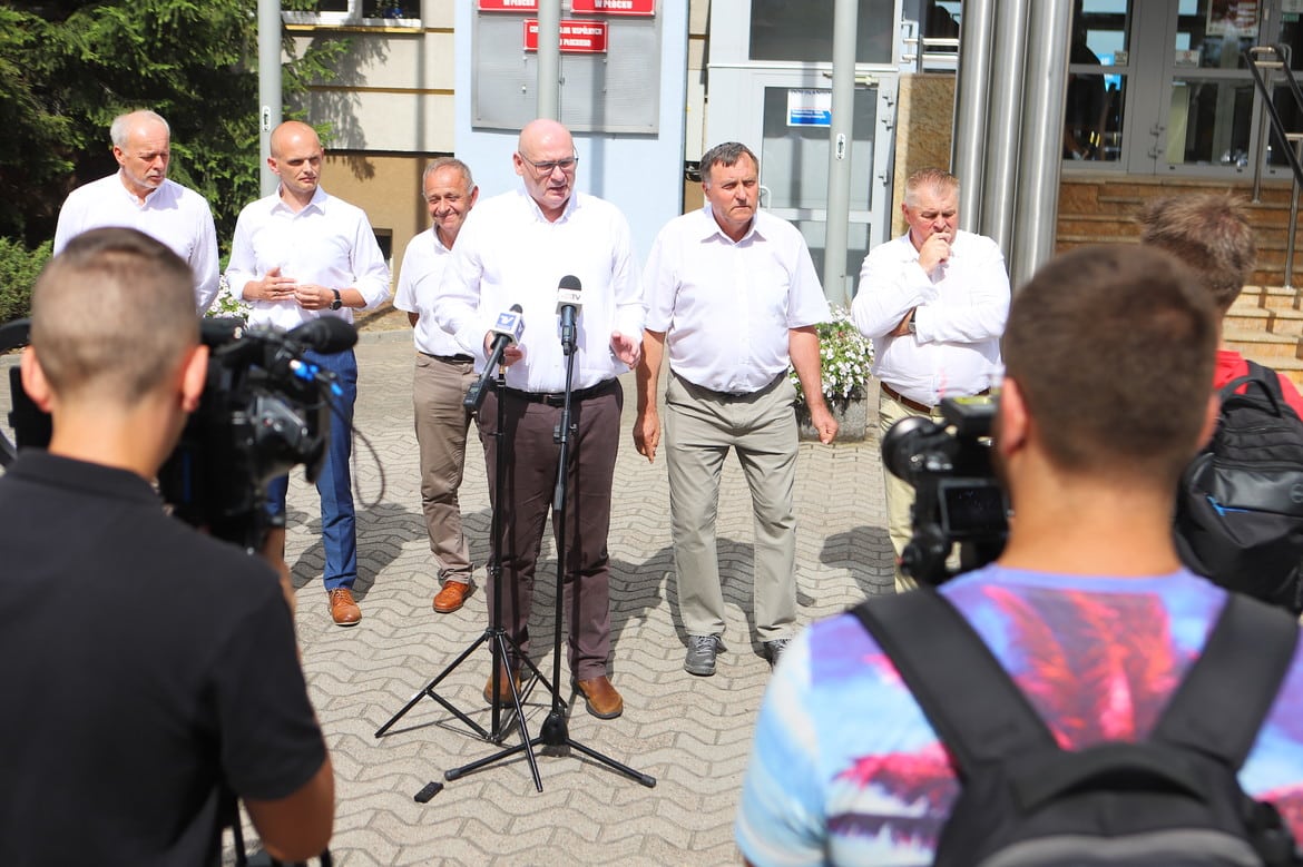 Na zdjęciu widać grupę mężczyzn w białych koszulach, którzy prowadzą konferencję prasową na zewnątrz budynku, otoczeni reporterami z kamerami.