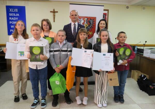 Na zdjęciu znajduje się grupa dzieci z dyplomami oraz odznaczeniami, które uczestniczyły w uroczystości wręczenia nagród, a w tle widoczne są elementy sali obrad.