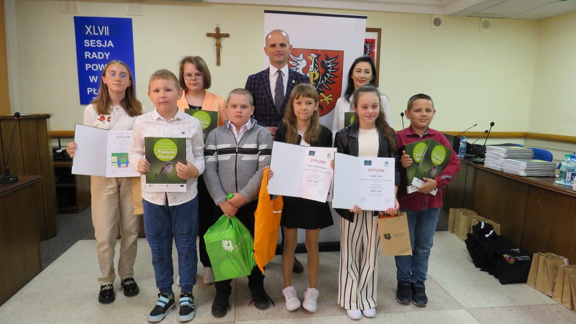 Na zdjęciu znajduje się grupa dzieci z dyplomami oraz odznaczeniami, które uczestniczyły w uroczystości wręczenia nagród, a w tle widoczne są elementy sali obrad.