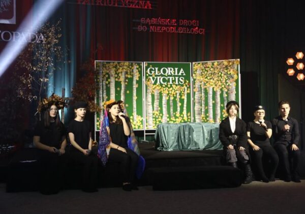 Na zdjęciu znajduje się grupa młodych artystów podczas występu na scenie, z elementami dekoracyjnymi w tle przedstawiającymi motyw przyrody oraz napis "GLORIA VICTIS".