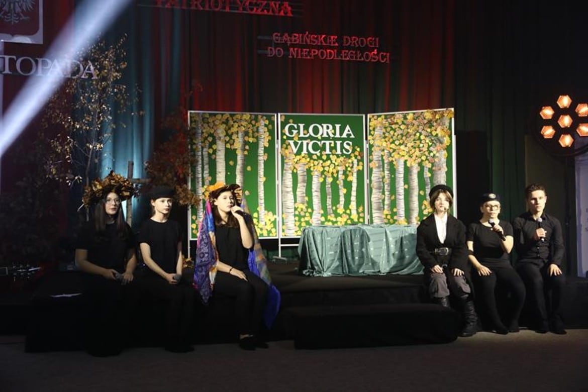Na zdjęciu znajduje się grupa młodych artystów podczas występu na scenie, z elementami dekoracyjnymi w tle przedstawiającymi motyw przyrody oraz napis "GLORIA VICTIS".