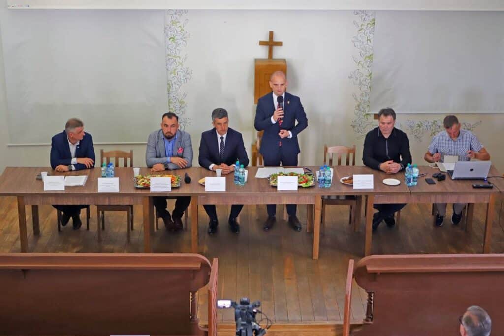 Na zdjęciu widoczna jest grupa mężczyzn siedzących przy stole podczas spotkania, gdzie jeden z uczestników przemawia, a na stole znajdują się napoje i jedzenie.