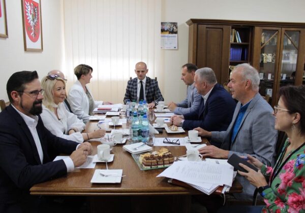 Na zdjęciu przedstawiono uczestników spotkania w sali konferencyjnej przy stole, gdzie omawiają sprawy przy filiżankach kawy i ciastach.