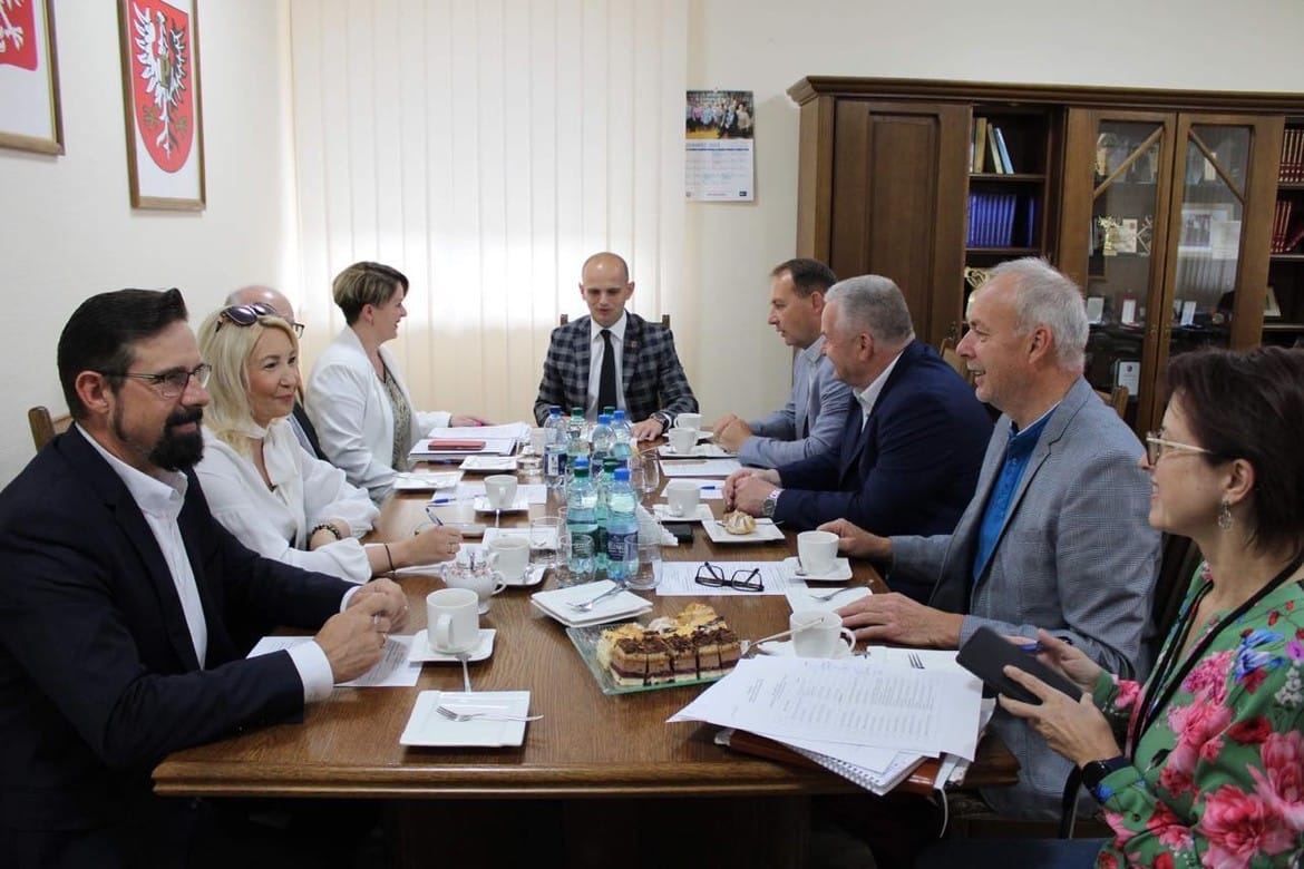 Na zdjęciu przedstawiono uczestników spotkania w sali konferencyjnej przy stole, gdzie omawiają sprawy przy filiżankach kawy i ciastach.