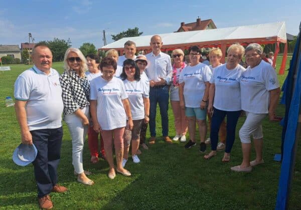 Grupa osób w koszulkach z napisem "Aktywny Senior" pozuje do zdjęcia na odkrytym terenie podczas wydarzenia społecznego.