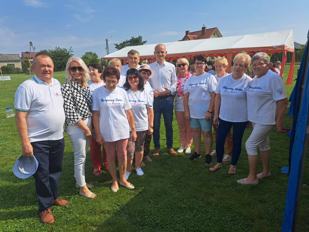 Grupa osób w koszulkach z napisem "Aktywny Senior" pozuje do zdjęcia na odkrytym terenie podczas wydarzenia społecznego.