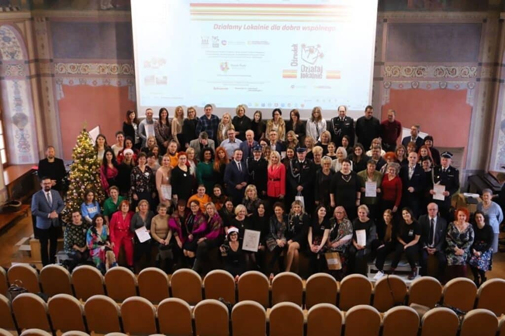 Na zdjęciu widać grupę ludzi zgromadzonych w sali, z choinką w tle, którzy trzymają certyfikaty, co sugeruje, że uczestniczyli w wydarzeniu związanym z tematyką lokalnej działalności.