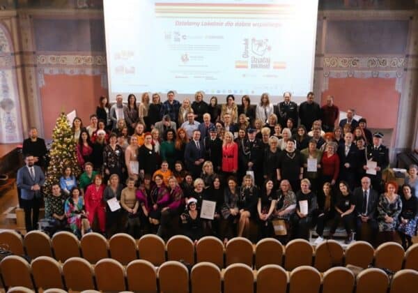 Na zdjęciu widać grupę ludzi zgromadzonych w sali, z choinką w tle, którzy trzymają certyfikaty, co sugeruje, że uczestniczyli w wydarzeniu związanym z tematyką lokalnej działalności.