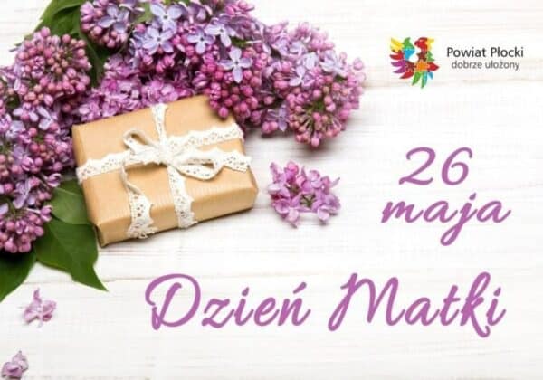 Zdjęcie przedstawia kwiaty bzu oraz zapakowany prezent z wstążką, na tle drewnianej powierzchni, z napisem "26 maja Dzień Matki".