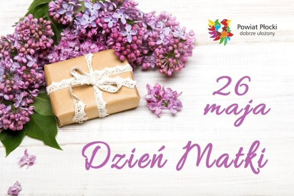 Zdjęcie przedstawia kwiaty bzu oraz zapakowany prezent z wstążką, na tle drewnianej powierzchni, z napisem "26 maja Dzień Matki".