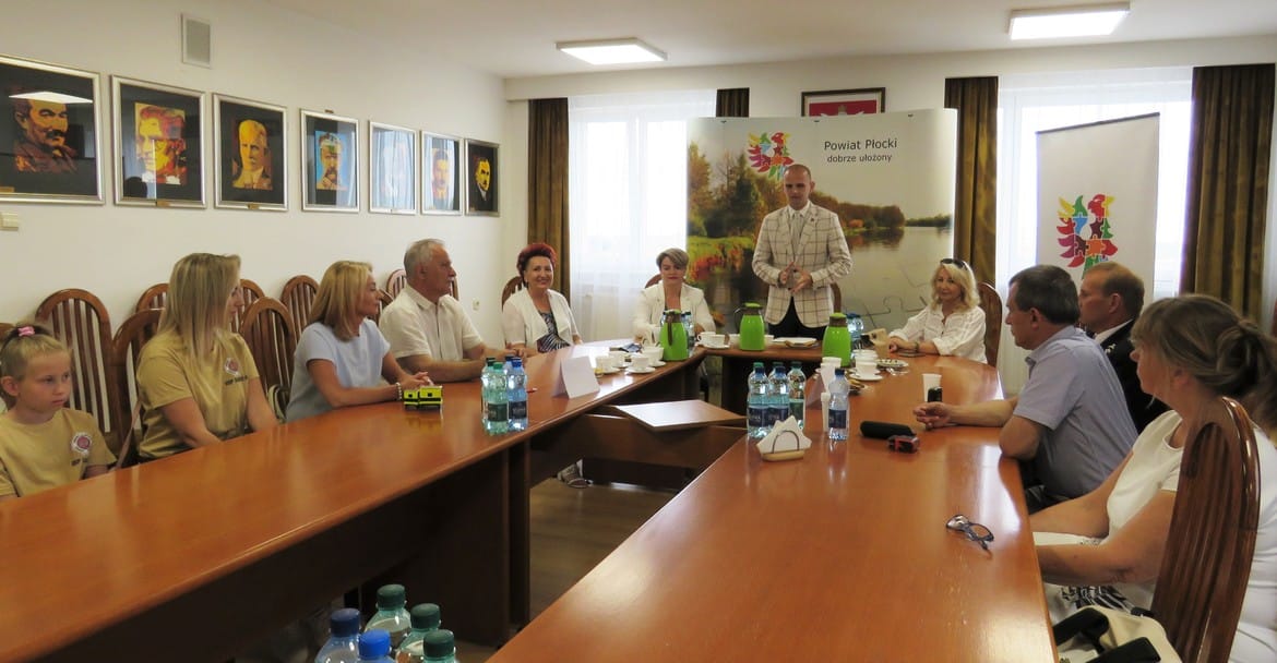Na zdjęciu widać grupę ludzi siedzących przy stoliku w sali konferencyjnej, podczas spotkania, z obrazami na ścianie w tle i banerem promocyjnym powiatu płockiego.