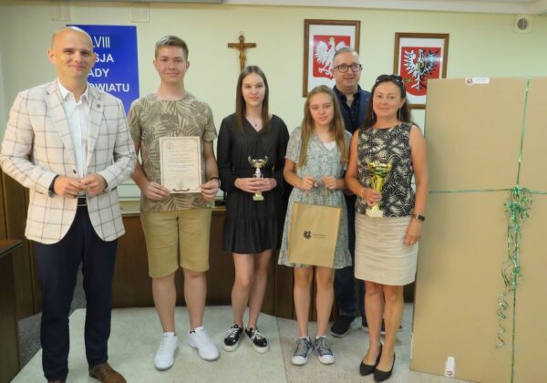 Uczestnicy uroczystości wręczenia nagród stoją razem w sali, trzymając puchary i dyplomy.